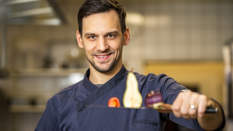 A cook in a blue uniform holds a knife with vegetables and fruit on it in a kitchen.