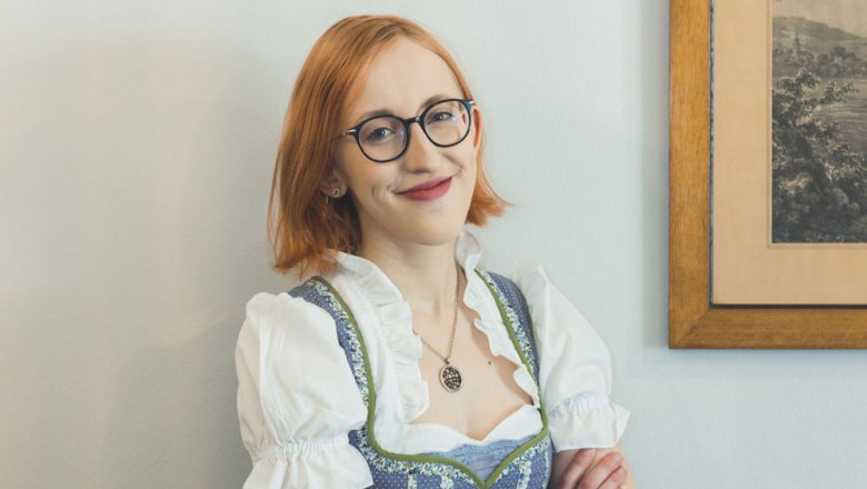 A woman with glasses and a dirndl smiles in front of a picture on the wall.