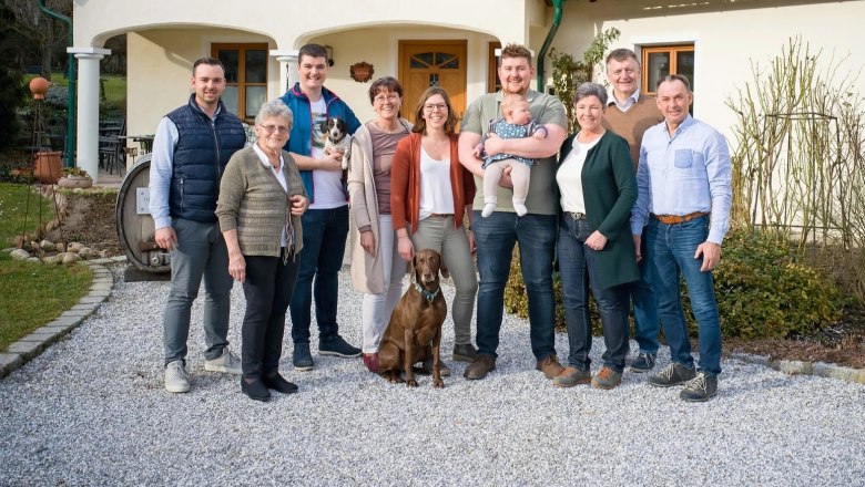 A family stands in front of a house, four generations together with a dog and a baby.