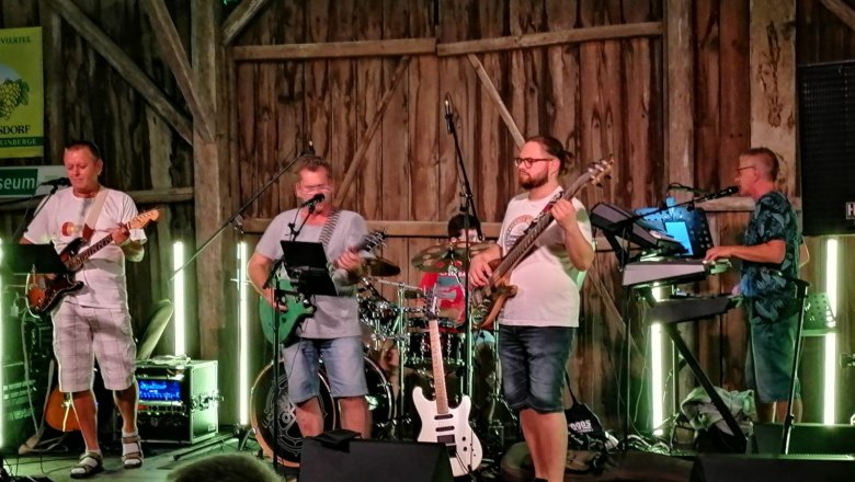 A band plays on a stage in a rustic wooden building. Five musicians with guitars, drums and keyboards can be seen.