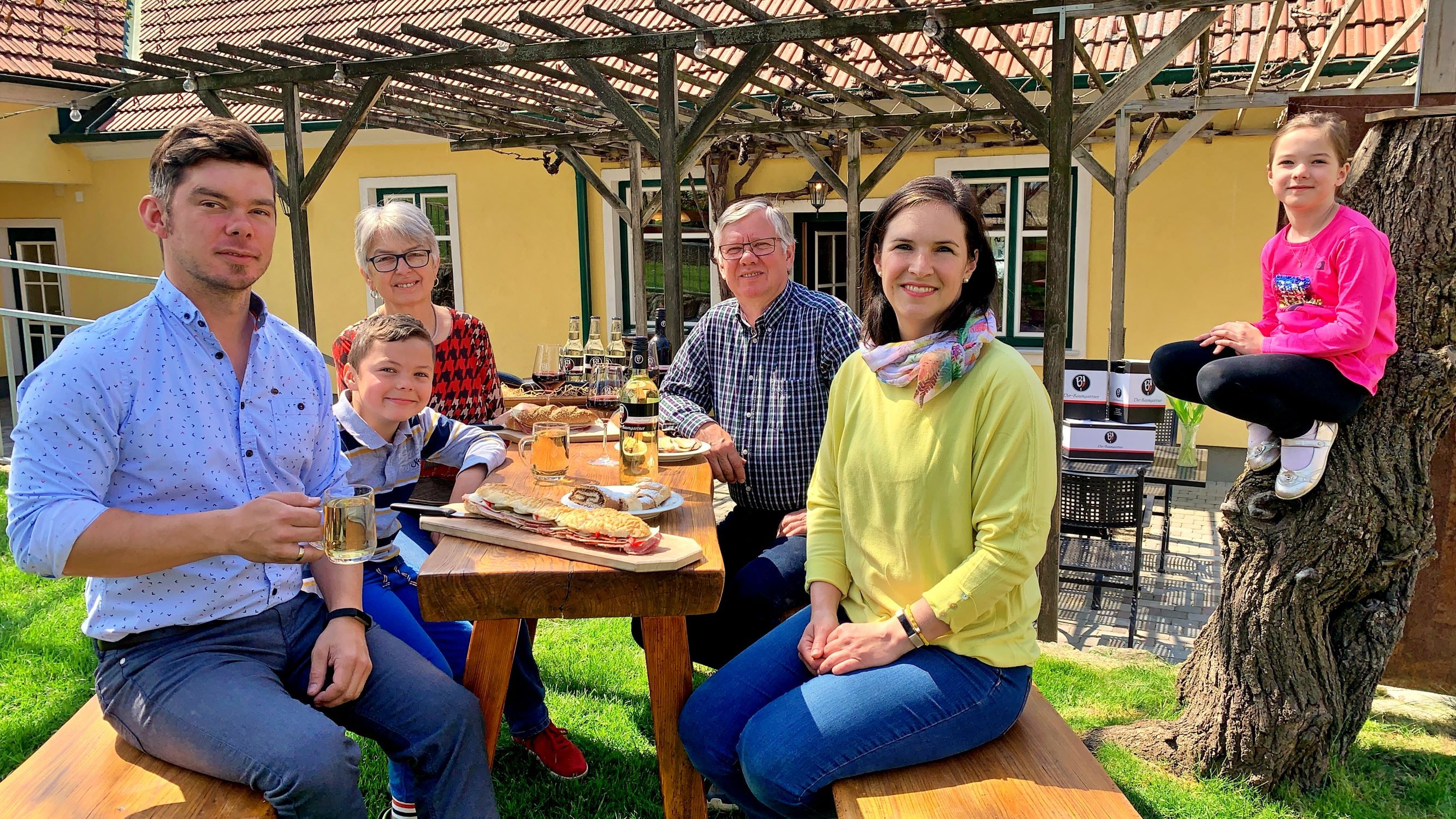 A family is sitting at a wooden table outside enjoying a meal. A child sits on a tree trunk. In the background is a yellow house with a pergola.