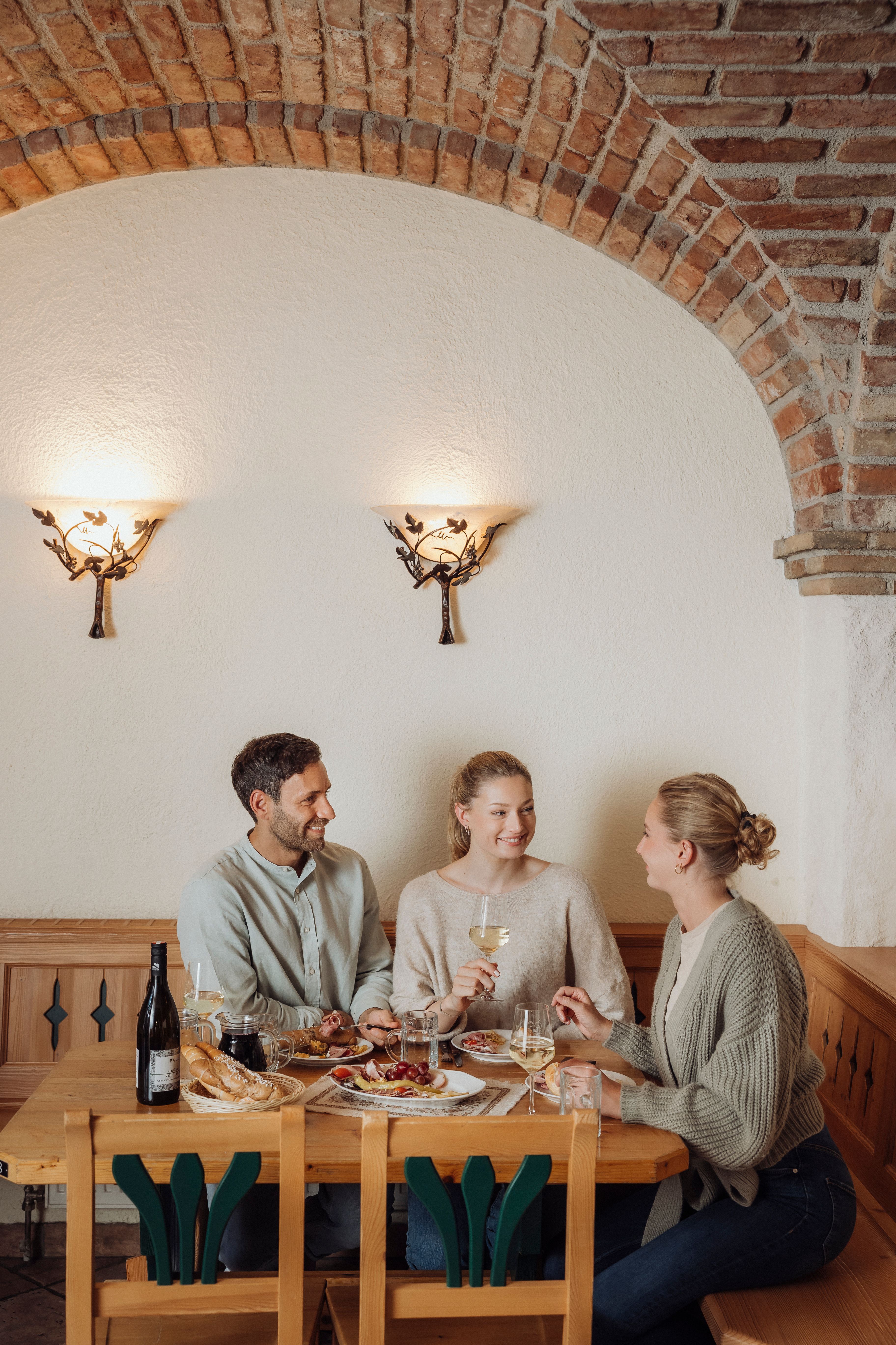 Three people sit at a wooden table in a wine cellar and enjoy wine and food.