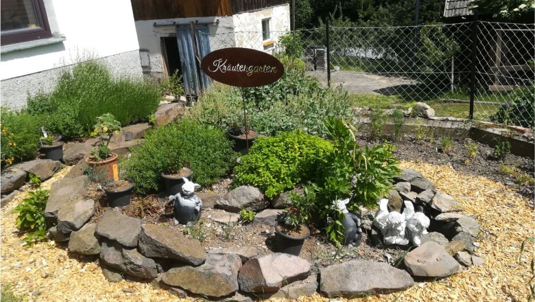 A herb garden with a stone border, plants and decorations, including garden figures. A sign with the inscription 'Herb garden' is visible.