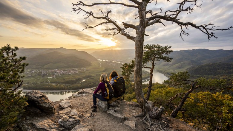 Two people sit on a rock and look out over a river landscape at sunset.