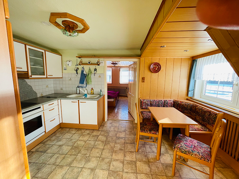 Cozy kitchen with corner bench, table and chairs, light wooden furniture, tiled floor, window with curtains, view into adjoining room.