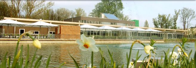 A modern building with a terrace and parasols by a pond with flowers in the foreground.