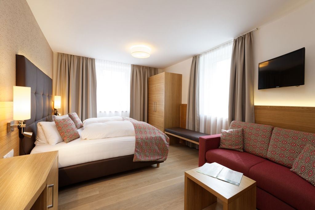 Modern hotel room with double bed, sofa, wardrobe and TV.