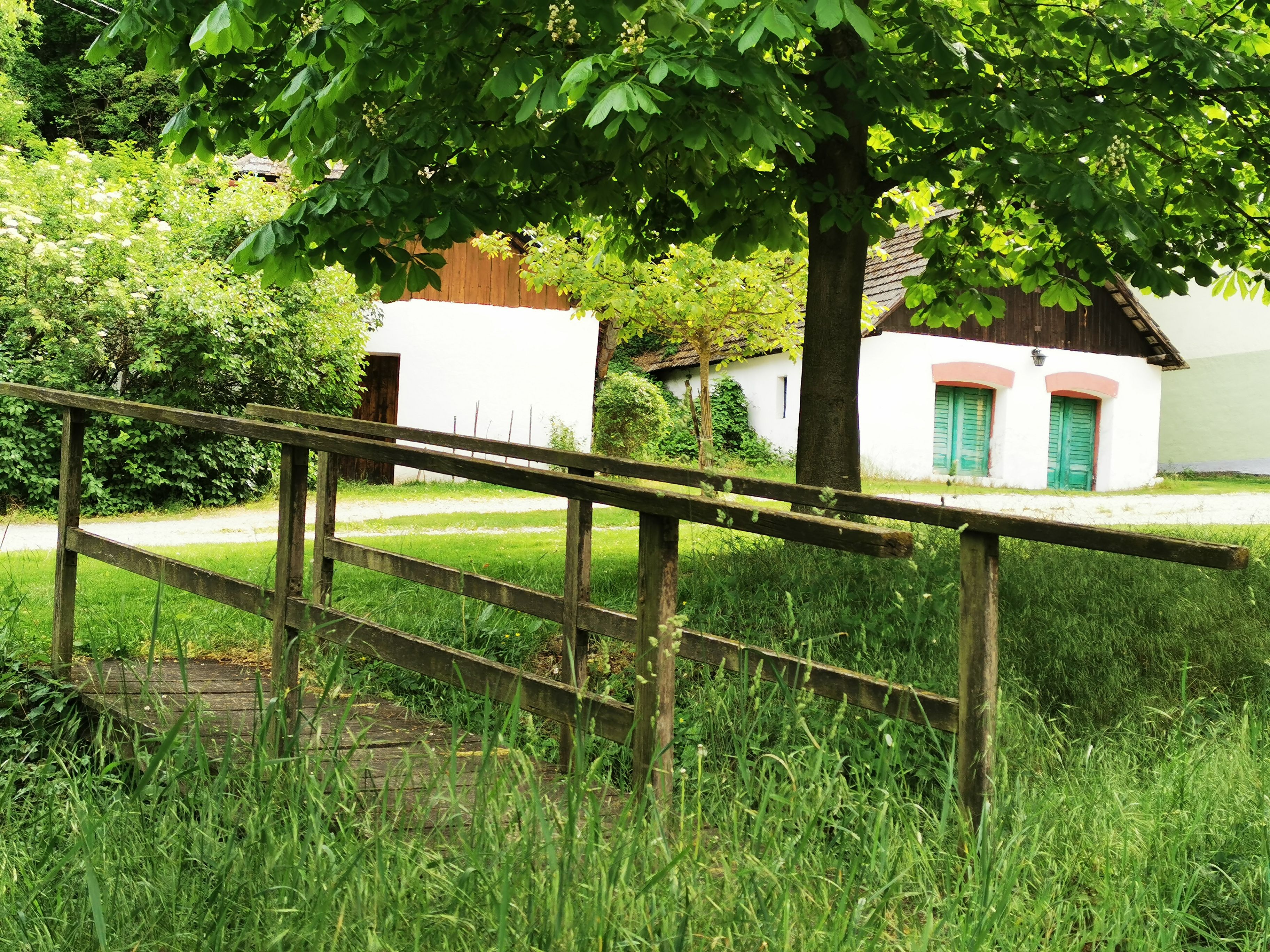 A small wooden bridge leads across a meadow to white buildings with green doors, surrounded by trees and bushes.