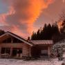 A wooden chalet in the snow at sunset with an orange sky and trees in the background.