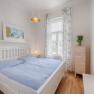 Bright bedroom with white bed, blue bed linen, wooden chest of drawers and window with curtains.