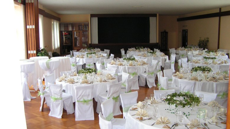 Elegant wedding hall with round tables, white tablecloths and chair covers, decorated with green plants and folded napkins.