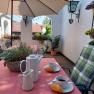 Cozy outdoor area with laid table, coffee cups and pastries, surrounded by flowers and plants.