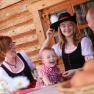 Family in traditional costume in front of a log cabin, laughing at the table.