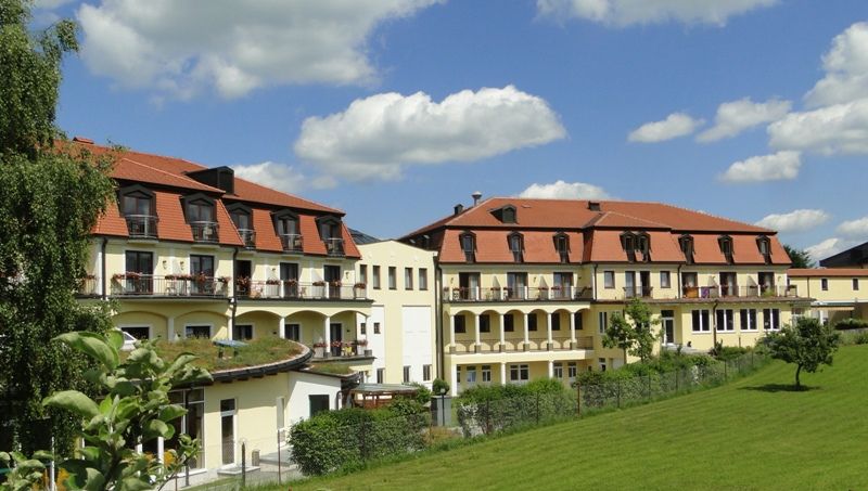 Kurhotel Moorbad Großpertholz with red roof and balconies, surrounded by green meadows and trees under a blue sky.