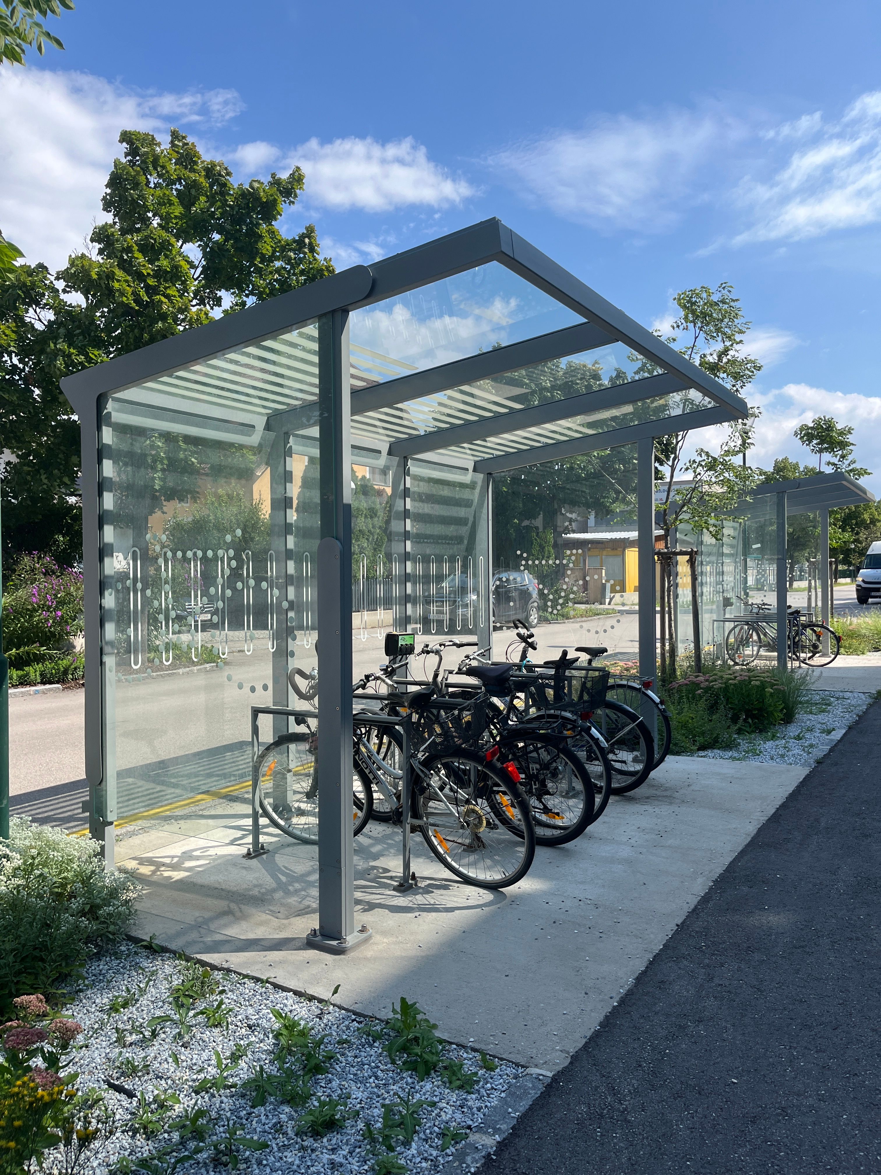Covered e-bike charging stations with several bicycles, surrounded by trees and plants.