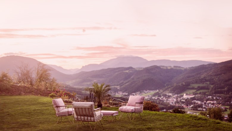 Distant view, © Matthias Kronfuss Garden furniture on a meadow with a view of a mountain landscape at sunset.