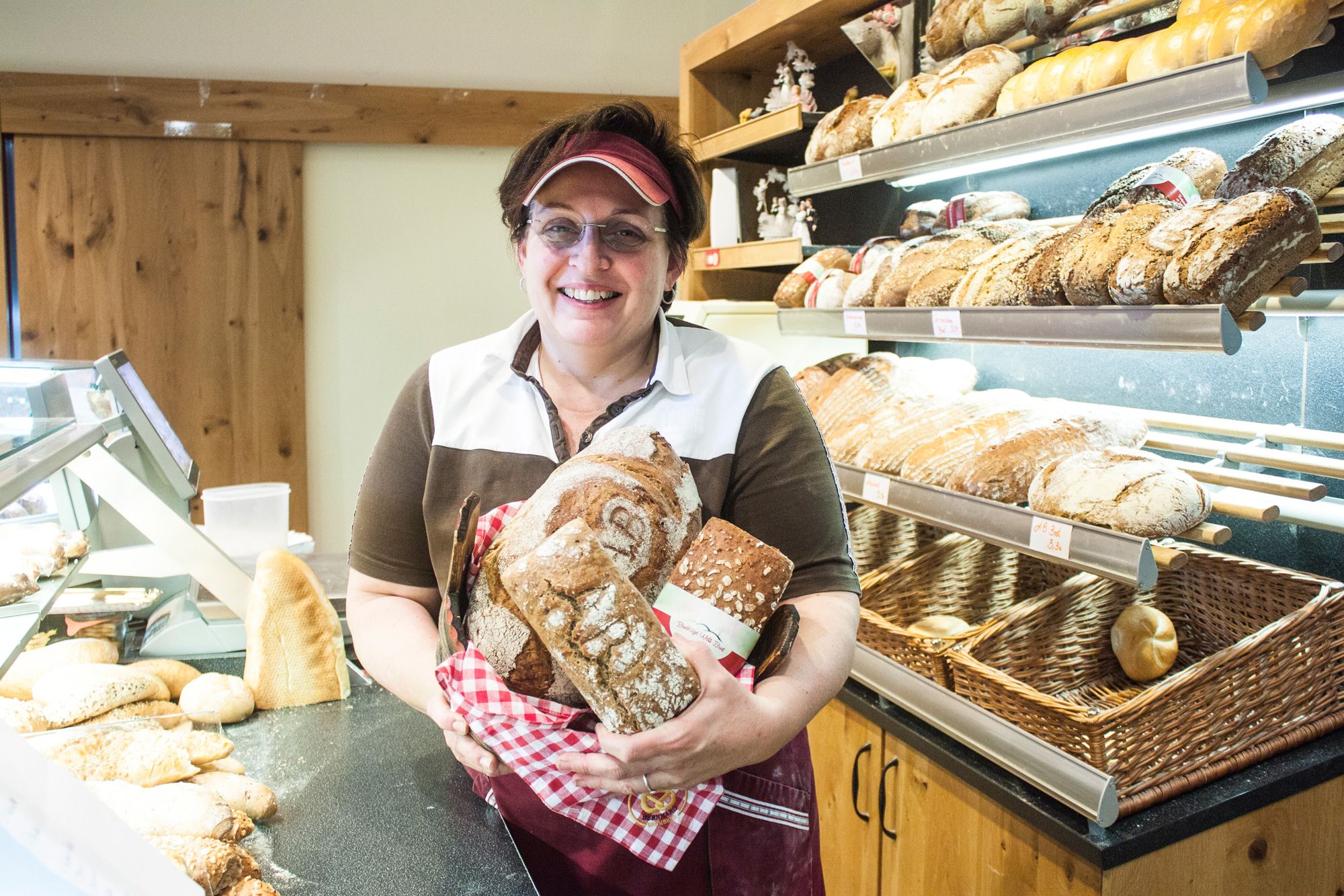 A woman in a bakery holds several loaves of bread in her hands and smiles at the camera.