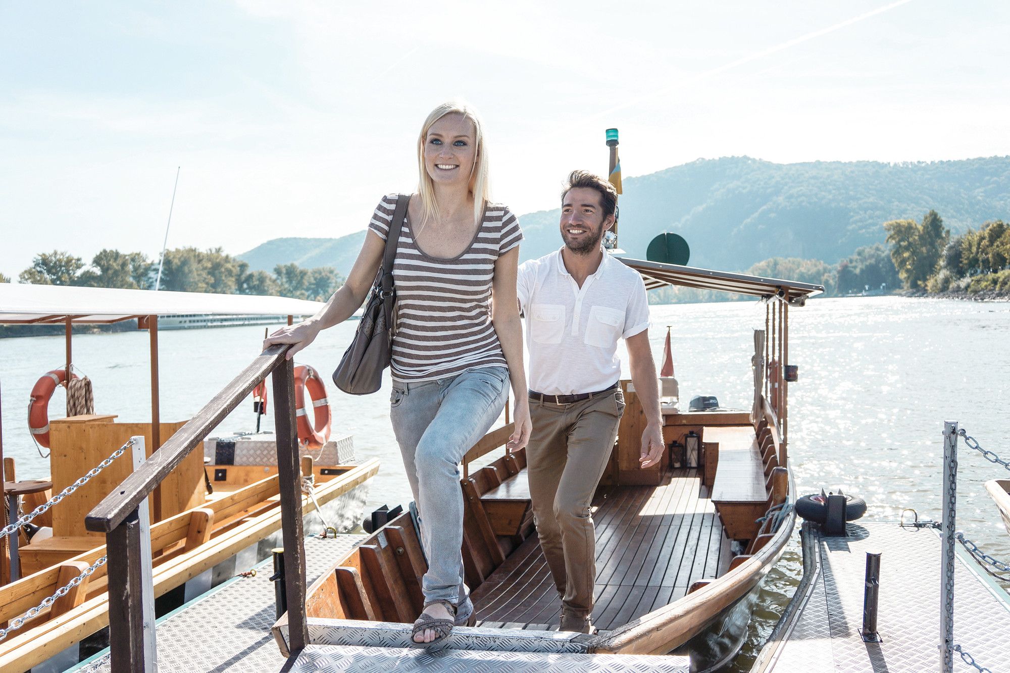 A man and a woman step off a boat onto a jetty. A river and wooded hills can be seen in the background.