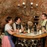 Four people in traditional dress are standing in a wine cellar holding wine glasses. They are laughing and chatting at a round table.