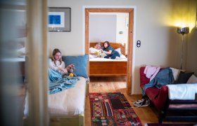 Interior view of a vacation apartment with two rooms. In the foreground a girl is sitting on a bed, in the background two people are reading on another bed.