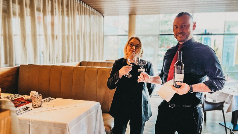 Two people in a restaurant toasting with wine glasses, one person holding a wine bottle.