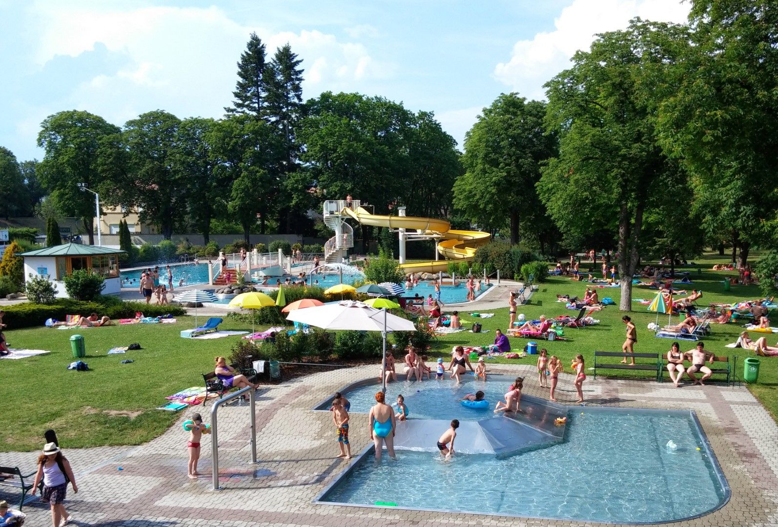 An outdoor pool with several pools, a yellow water slide and lots of people relaxing on the lawn.
