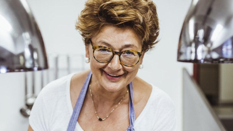 A smiling woman with glasses and an apron in a kitchen.