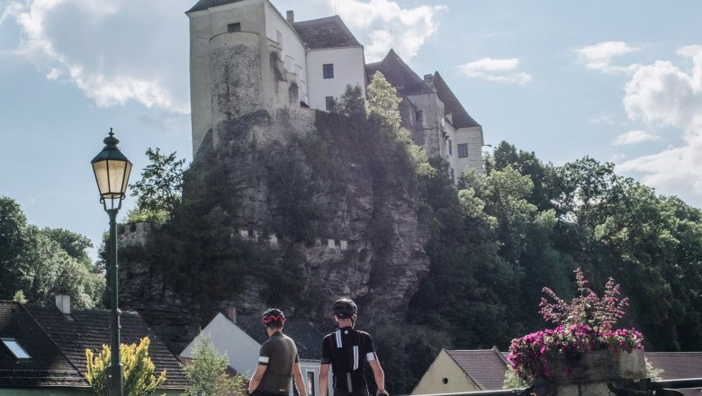 Two cyclists are standing on a bridge with a view of Raab Castle, which is enthroned on a rock.