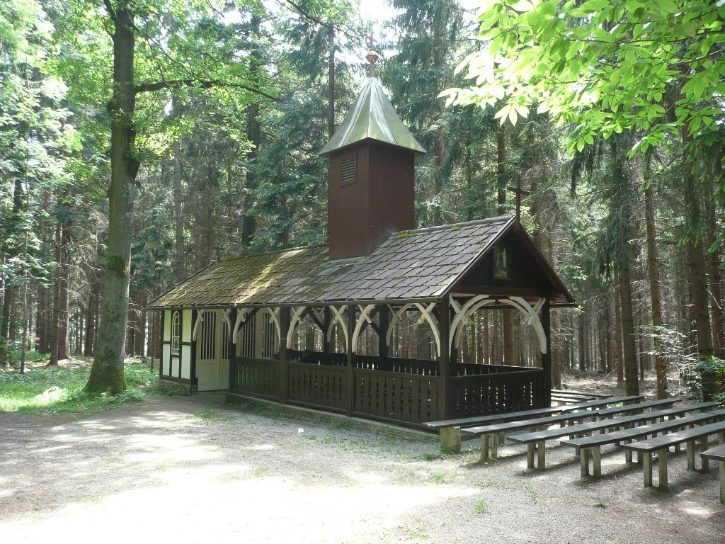 A small chapel in the forest with wooden benches outside.