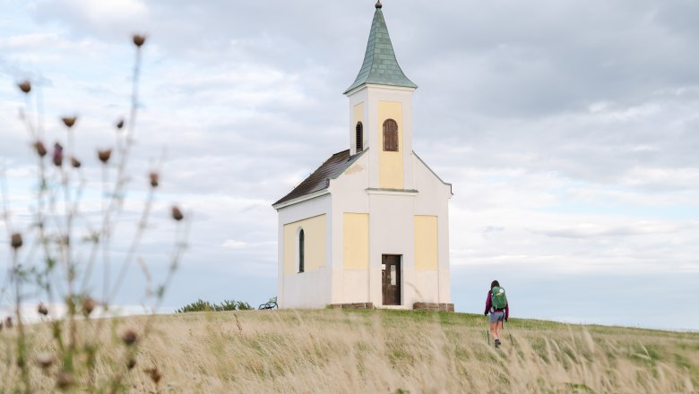 A small chapel on a hill with a person walking towards it, surrounded by a meadow and a cloudy sky.