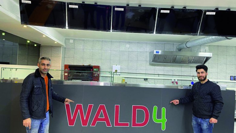Two men are standing in front of a counter labeled 'WALD4' in a pizzeria.