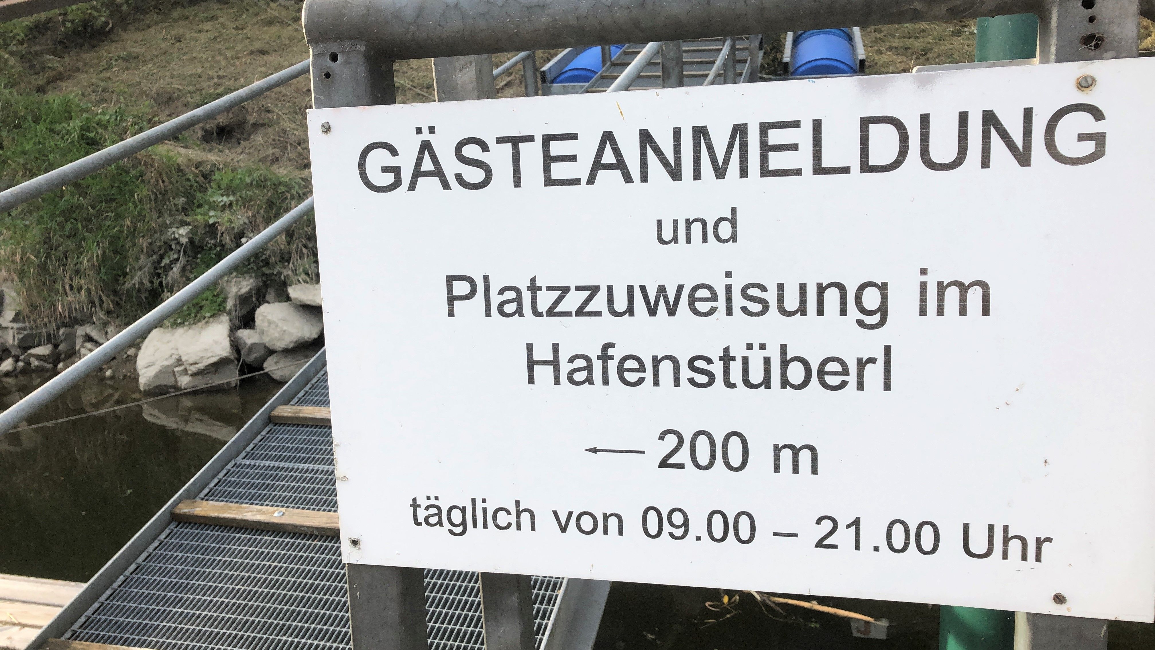 A sign with the inscription 'Gästeanmeldung und Platzzuweisung im Hafenstüberl, 200 m, täglich von 09.00 - 21.00 Uhr'.