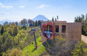 Wooden building with heart window and cable car on a wooded hill, mountain in the background.
