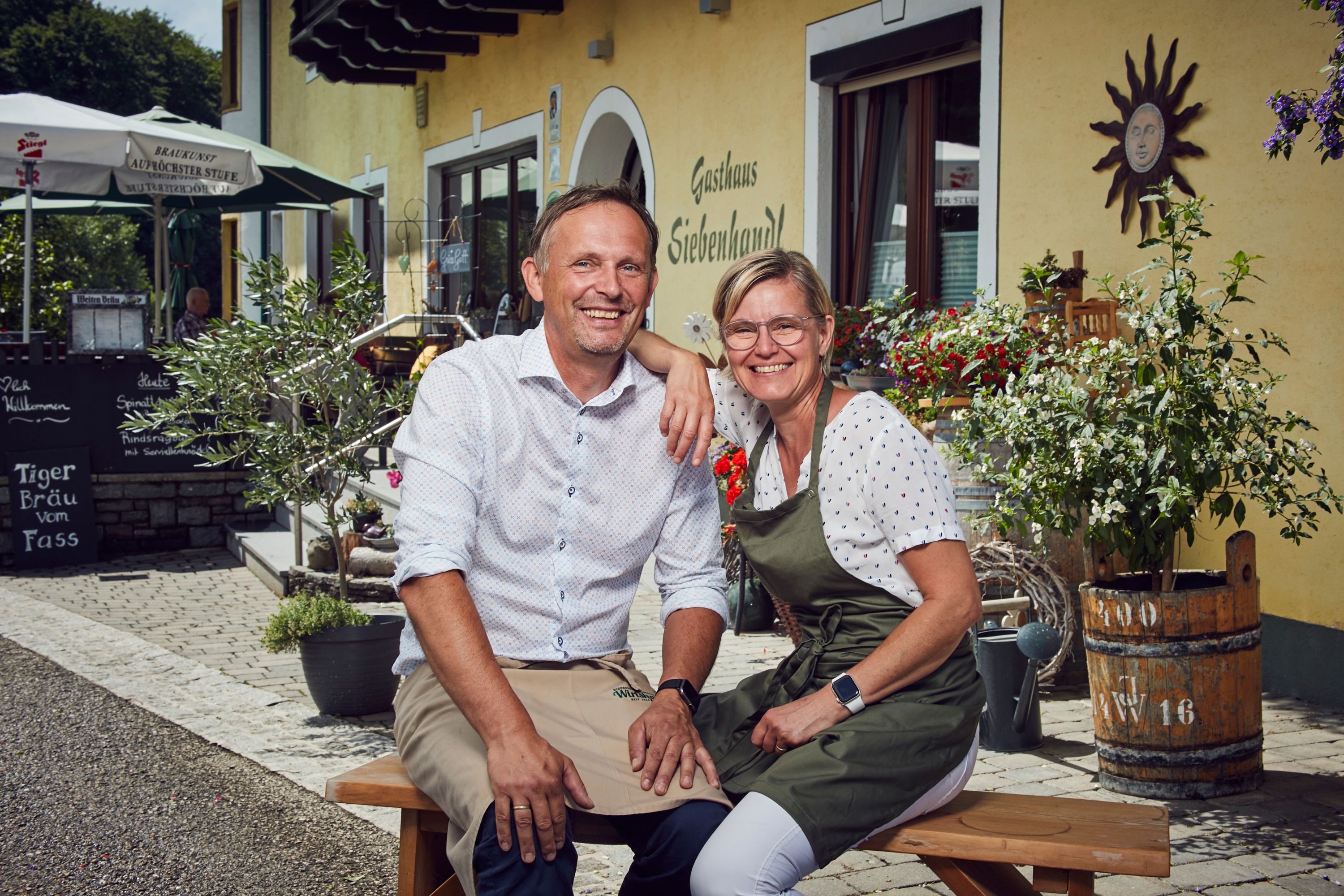 Smiling couple in front of Gasthaus Siebenhandl, surrounded by plants and decoration.