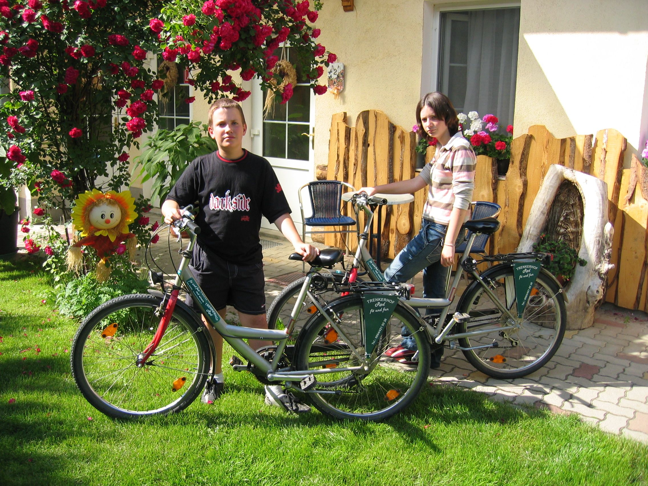 Two people with bicycles in front of a house with flowers and wooden decorations.
