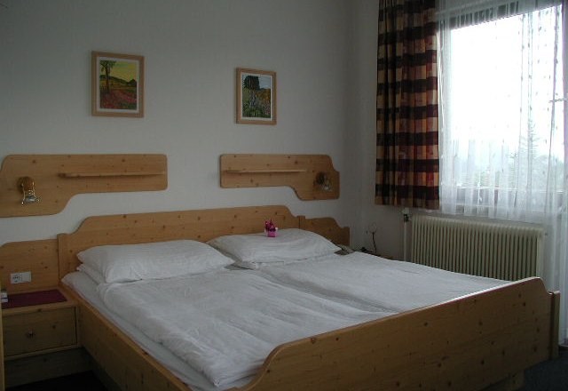 A simply furnished hotel room with a wooden double bed, two pictures on the wall and a window with curtains.