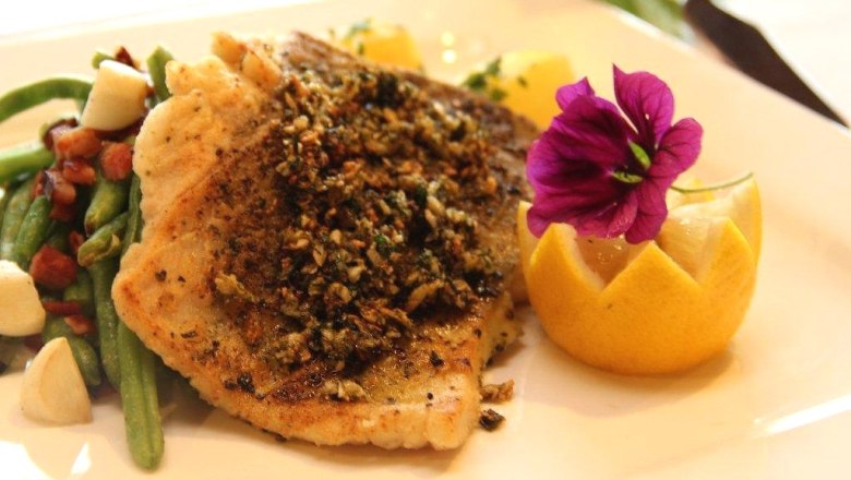 A plate of fried fish fillet, decorated with a flower and a slice of lemon, served with green beans and bacon.