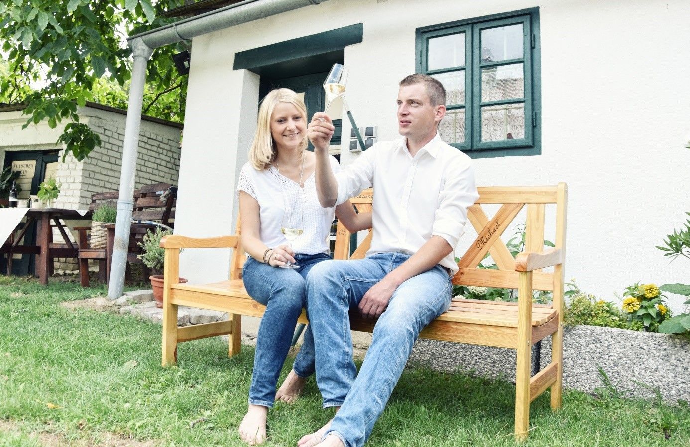 A couple is sitting on a wooden bench in front of a house, clinking glasses of wine.