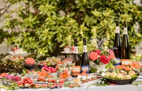 A richly laid outdoor table with bottles of wine, glasses, flowers and various dishes.