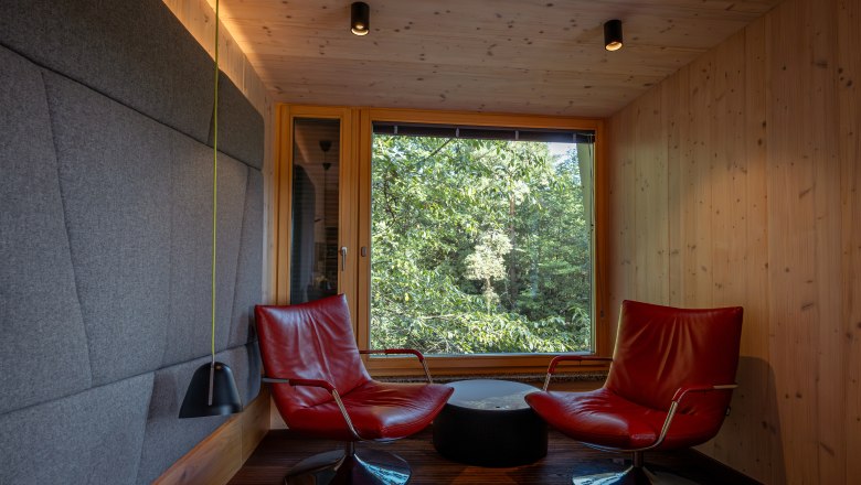 Interior view of a modern tree house lodge with two red armchairs in front of a large window with a view of the forest.