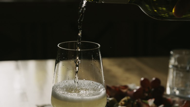 A glass of white wine is poured at a table with a cheese and cold meat platter in the background.