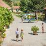 A sunny courtyard with people walking, playing table tennis and chatting. Surrounded by plants and buildings with red roofs.