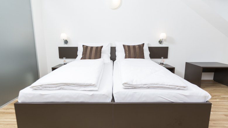 Modern guest room with two single beds, white comforters and brown pillows, wooden floor and bedside tables with lamps.