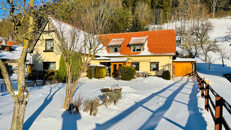 Snow-covered house with red roof and garden, surrounded by trees and fence, in sunny weather.