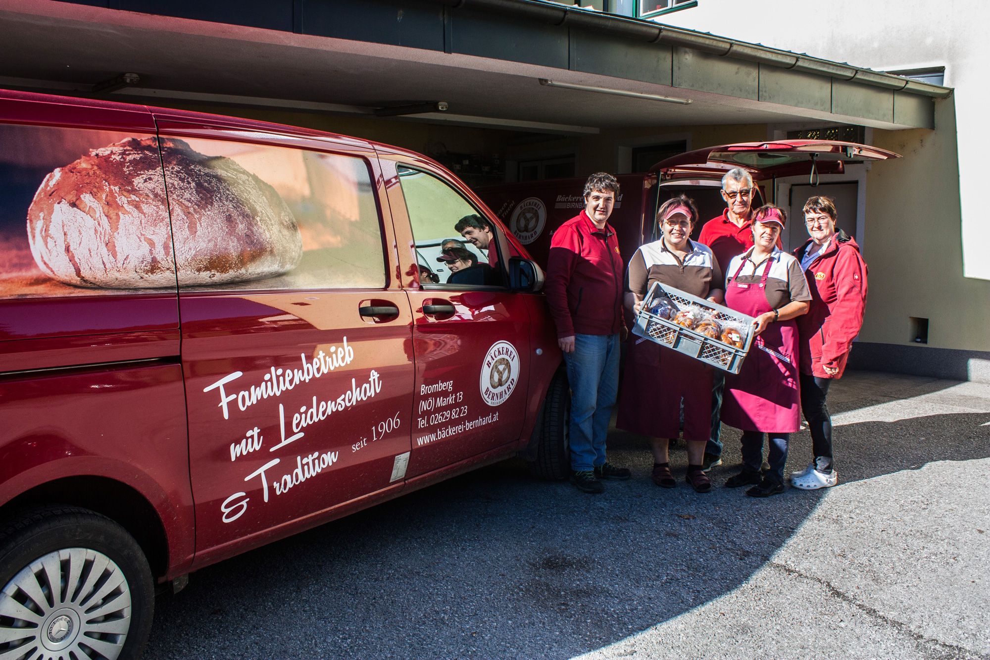 Five people hold a gray box of pastries next to a red van.