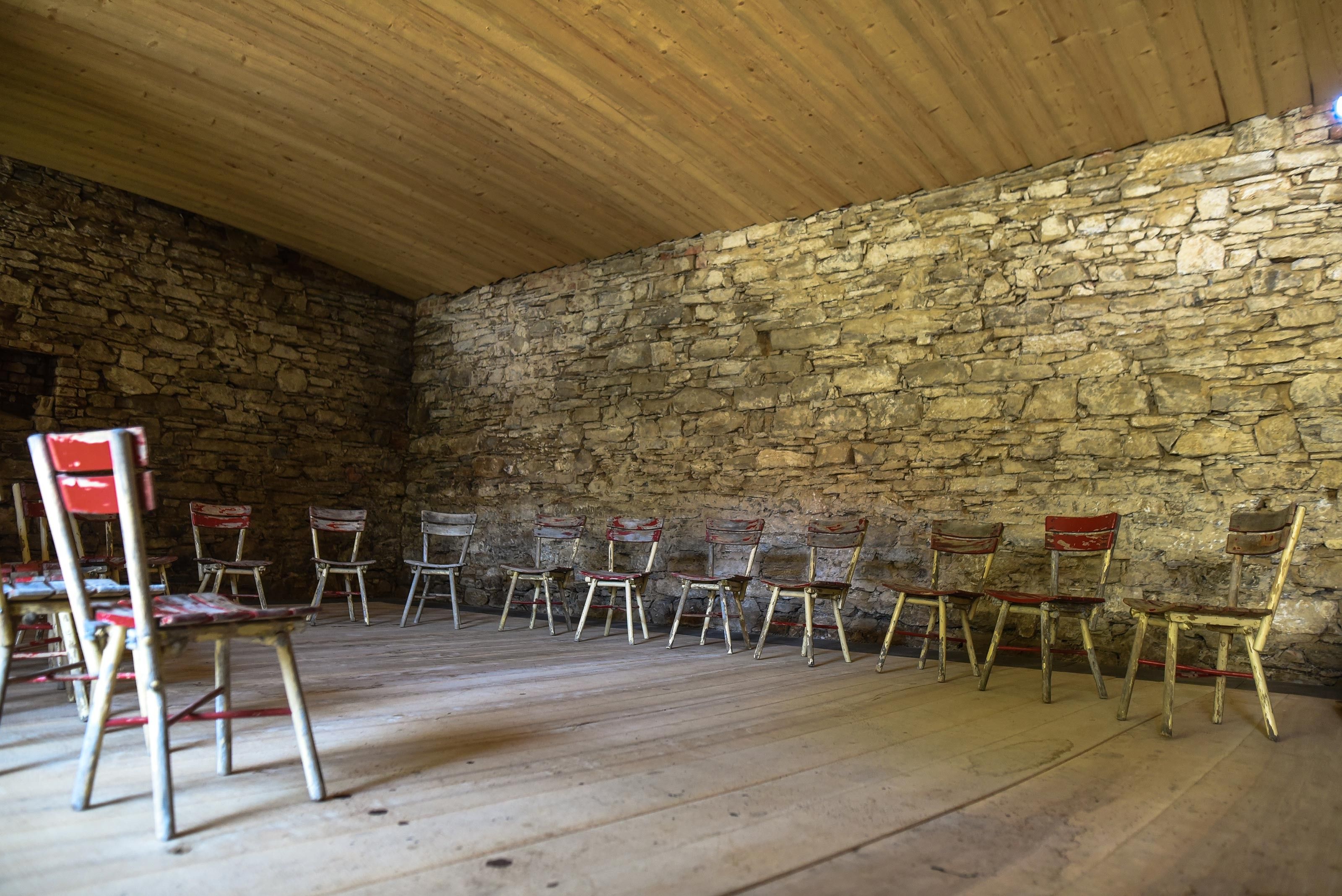 An empty room with a stone wall and a wooden ceiling. Several old, colorful chairs are arranged in a semicircle.