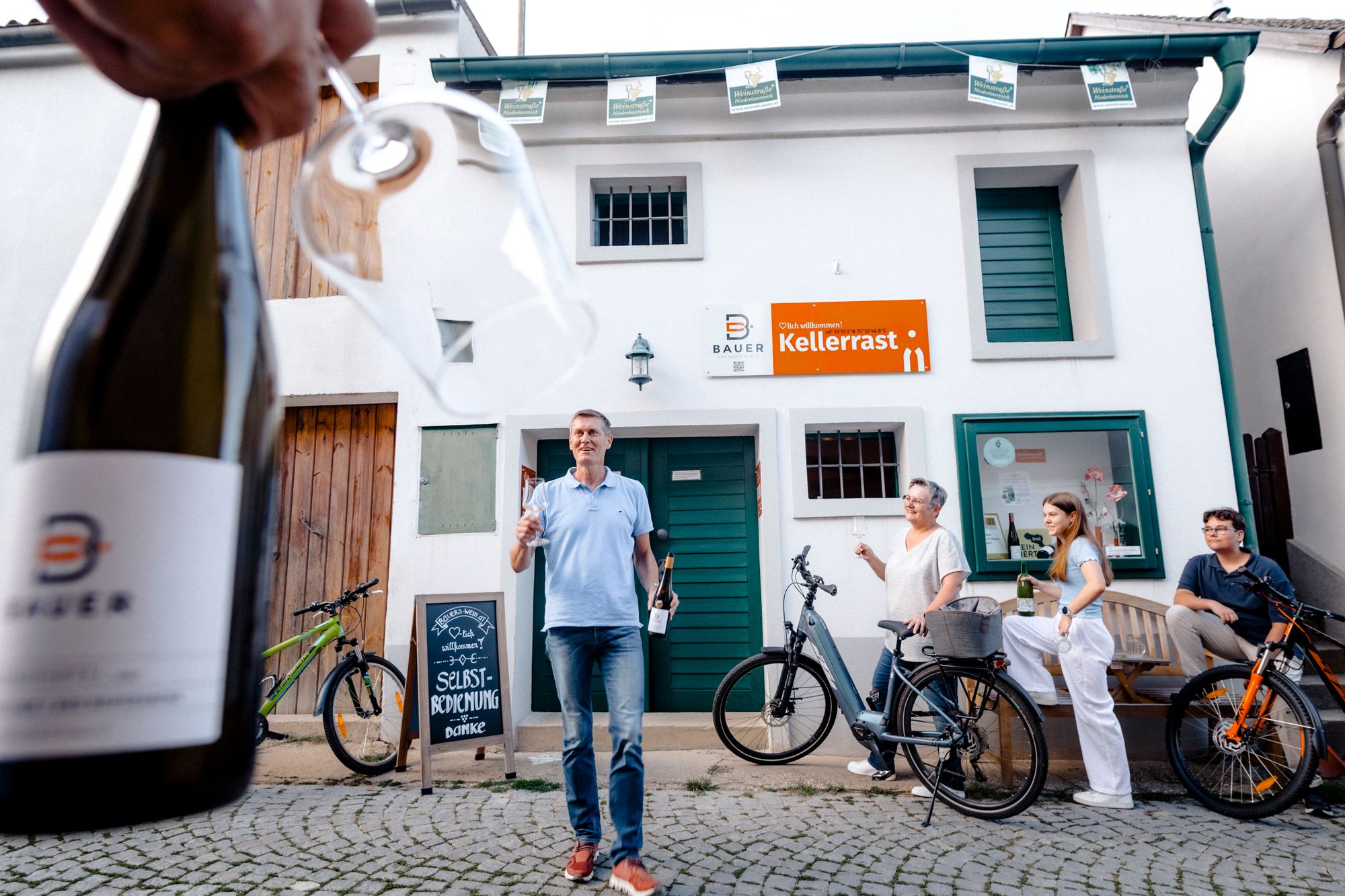 People stand in front of a building with a sign 'Kellerrast' (cellar rest), while a person in the foreground holds a bottle of wine and a glass.
