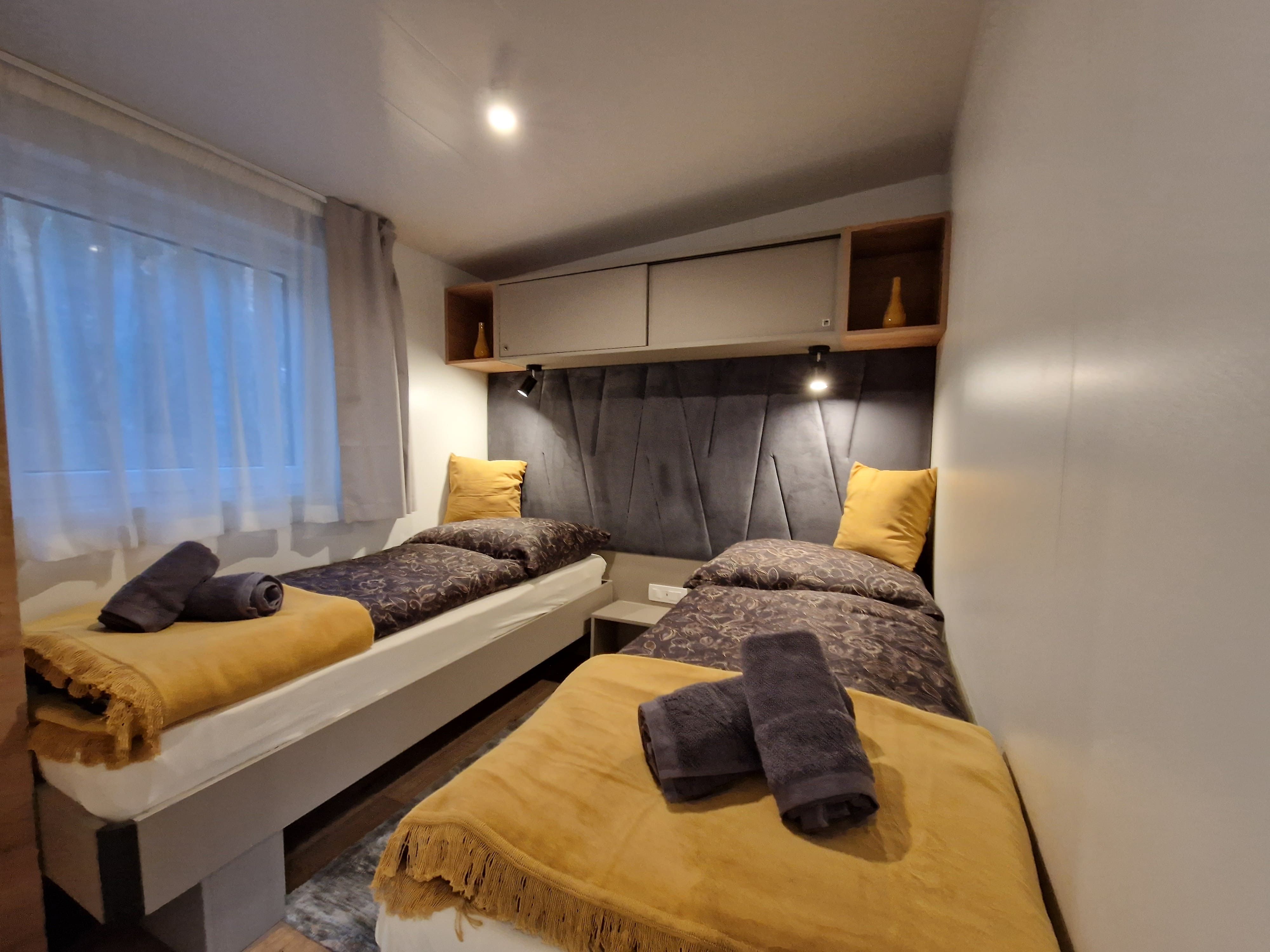 Two single beds in a cozy chalet room with yellow blankets and pillows, grey towels and a window with a curtain.