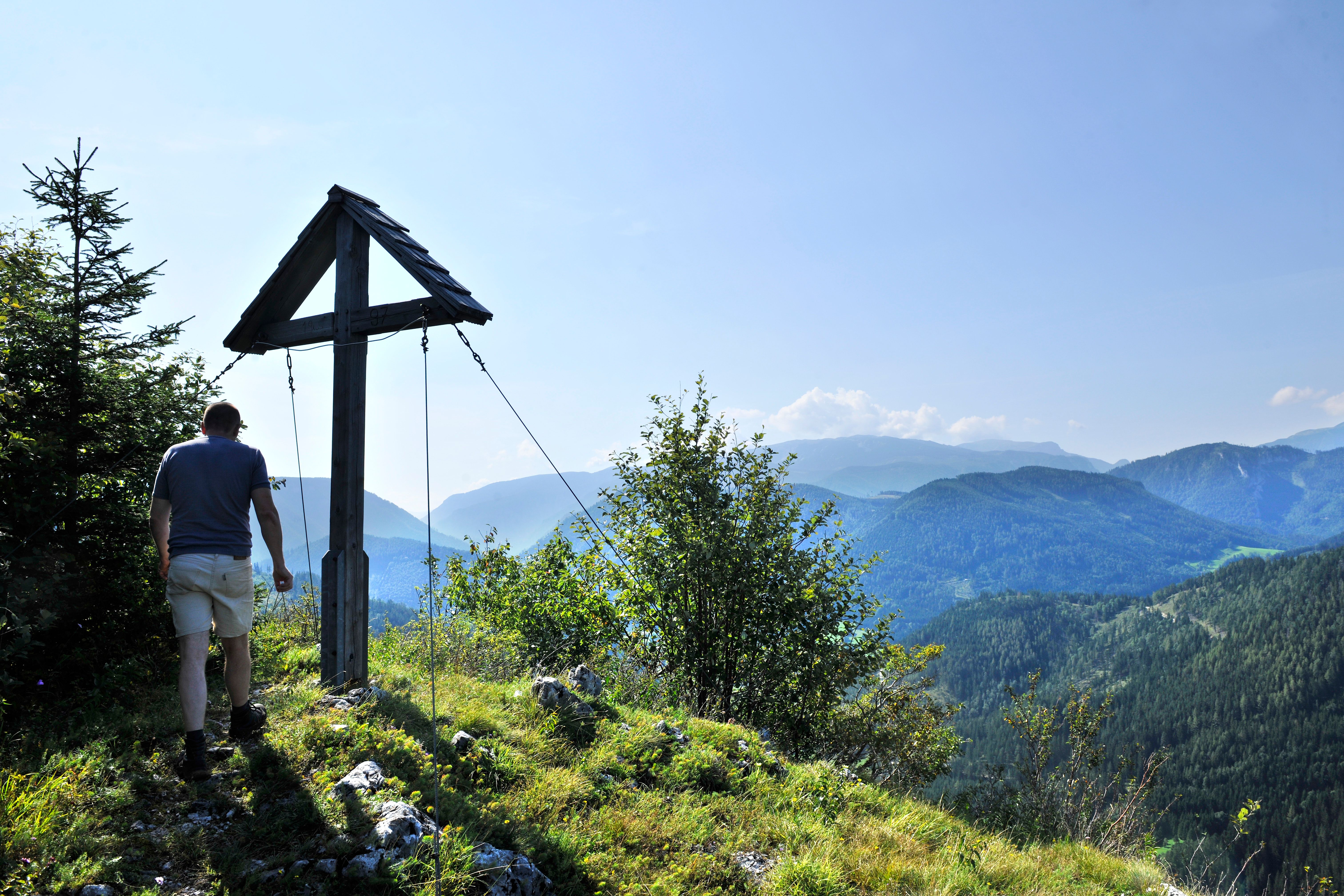 A hiker stands next to a summit cross with a view of wooded mountains.