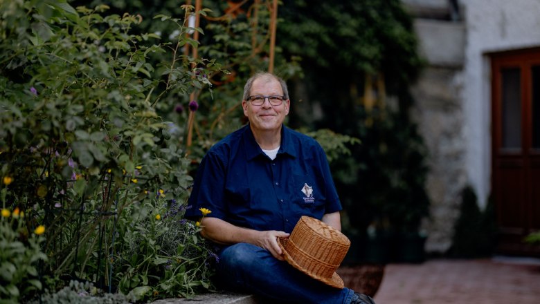 A man sits in a garden and holds a woven hat.